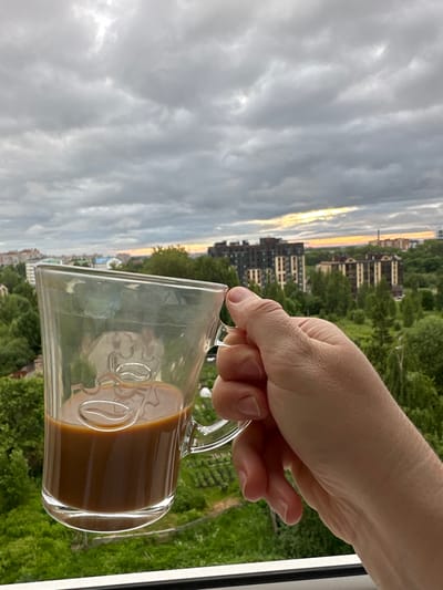 A hand holds a clear glass mug filled with coffee against a cloudy sky and an urban landscape. The mug has embossed coffee bean designs. In the background, apartment buildings and green trees are visible. The sky is overcast with hints of sunlight breaking through near the horizon. The scene appears to be a residential area in Rubtsovo, Russia.