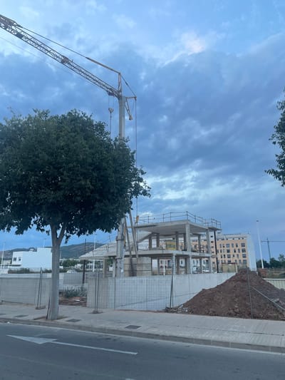The image depicts a construction site in Sagunto, Spain, under a cloudy sky. A large, dark green tree dominates the foreground, its dense foliage obscuring parts of the scene. To the left, a yellow crane extends upwards, its arm reaching towards the sky. Behind the tree and crane, a multi-story building under construction is visible, with its concrete framework exposed and scaffolding attached. Further in the background, a taller, light-colored building with many windows stands, suggesting an established urban area. A pile of dark soil is situated near a chain-link fence, separating the construction from a paved sidewalk and a road. The road has a white arrow painted on it, indicating a direction of traffic. The overall impression is one of ongoing development within a coastal or Mediterranean setting, with the clouds suggesting a slightly overcast day, possibly in the late afternoon or early morning. No people are visible in the image, and no text is discernible.