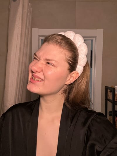 The image depicts an indoor scene in the city of Nikol'skii, Russia. A light-skinned female individual, with blonde hair pulled back into a ponytail, is positioned centrally. She wears a white, puffy-textured headband and a black, satin-like robe. Her facial expression is a pronounced grimace, characterized by tightly squinted eyes, a deeply wrinkled nose, and an open mouth revealing gold-colored dental braces on her upper teeth. To the left of the subject, a light-colored vertical curtain is visible. Behind her, a white doorframe with molding is present. To the right, a dark, multi-shelf storage unit is partially visible, containing several light-colored objects, possibly baskets or containers. The scene is illuminated by warm, artificial light.