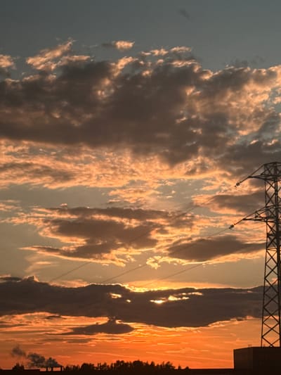 This landscape image captures a striking sunset or sunrise, characterized by a dynamic sky filled with large, illuminated clouds and the silhouette of a power line tower. The scene is dominated by a vast expanse of sky, rich with warm, golden, and orange hues emanating from the low-hanging sun, which is partially obscured by dense cloud formations towards the right side of the frame.

Numerous voluminous clouds, appearing to be cumuliform, are dramatically lit from behind and below by the sun's intense light. Their edges glow with bright oranges and yellows, contrasting with their darker, shadowed undersides, which display shades of grey and purple. This lighting creates significant depth and texture within the cloudscape. The sky transitions from these warm tones near the horizon and sun to a cooler, muted blue-grey in the upper and far-left portions of the image.

Running across the upper-middle section of the frame is the prominent silhouette of a metal lattice-work power line tower. Several power lines extend from this tower, appearing as thin, dark strands against the bright sky and illuminated clouds. On the distant horizon at the very bottom of the image, a dark, silhouetted band of trees is visible, along with some indistinct man-made structures or buildings on the far right.

No people or discernible activities are visible, emphasizing the natural beauty of the celestial event. The time of day is clearly either dawn or dusk, lending a serene yet dramatic atmosphere to the scene. The presence of the power lines suggests an area where natural landscapes meet human infrastructure, possibly on the outskirts of a town or rural-industrial fringe. No visible text is present in the image.