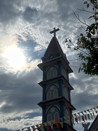The image shows the bell tower of a church, likely belonging to a Catholic church, in Suối Tân, Vietnam. The tower is tall and slender, with a steeply pitched, tiled roof. It features multiple levels, each with arched windows or decorative elements. A prominent cross is visible at the very top of the tower. In the foreground, strings of colorful triangular flags are strung across the lower part of the frame, suggesting a festive occasion. To the right of the tower, branches of a leafy tree extend into the frame. The sky is dramatic, with heavy, dark clouds interspersed with areas of bright sunlight, creating a striking contrast and a halo effect around the sun. The scene is captured from a low angle, looking upwards towards the sky and the church tower.