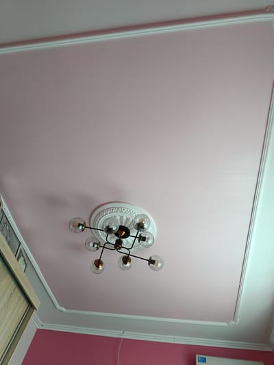 A medium shot captures a modern chandelier with clear glass globe shades, each housing a filament bulb, suspended from a pink ceiling. The chandelier is mounted on a decorative white ceiling medallion. The ceiling itself is framed by white crown molding, which transitions to a white inset panel on the right side of the frame, and on the left, a portion of a wooden wardrobe is visible. The wall below the crown molding is painted pink. The scene suggests an indoor setting, likely a home, with the lighting from the chandelier casting shadows on the ceiling. The overall impression is one of domestic interior design. There are no people visible, nor any discernible weather or time of day cues beyond what is suggested by indoor lighting. No text is visible in the image.
