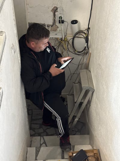 The image displays an indoor scene within a narrow stairwell or utility area in Kaloyanovo, Bulgaria. One adult male, with short dark hair and a sturdy build, is positioned on a tiled stair, leaning slightly against the left wall. He wears a black jacket with a red zipper and an "A1" logo on the left chest, black athletic pants with three white stripes, and dark athletic shoes with red accents. He is holding a black digital tablet or large smartphone with both hands, focusing on its illuminated screen.
Mounted on the right wall, above the male's eye level, is network equipment, including a white rectangular Wi-Fi router with multiple antennas and a white cylindrical device. Numerous yellow and black cables are present, with a large coil of black cable secured to the wall and other wires exposed around a wall-mounted power outlet. A silver, partially folded four-step ladder rests against the lower section of the right wall. On the tiled floor in the bottom right corner, an open brown cardboard box contains a black network device and coiled cables. The walls are light-colored, textured, and exhibit visible imperfections. The floor consists of light-toned tiles interspersed with dark, patterned sections.