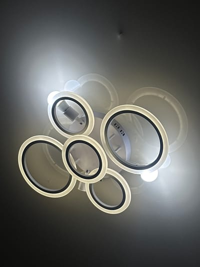 The image depicts a modern, abstract ceiling light fixture composed of several interconnected oval rings. The rings are illuminated from within, casting a bright, diffused light. The fixture is mounted on a plain, grey ceiling, which appears to have a slight texture. The lighting creates soft shadows and highlights on the ceiling, giving the fixture a somewhat ethereal quality. The overall impression is one of contemporary design and understated elegance. The location of this image is unknown, and no details about Nookat, Kyrgyzstan are present in the visual information provided.