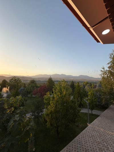 The image depicts a scenic view of a park or garden area, likely in Karankultugay, Uzbekistan, during what appears to be late afternoon or early evening, given the golden hue of the sky and the long shadows. In the foreground, a tiled patio or walkway extends into the frame, with a portion of a building's overhang visible at the top right. This overhang features a modern light fixture.
The middle ground is dominated by lush greenery, with various trees of different sizes and types, including evergreens and deciduous trees with vibrant red foliage. A fountain with water spray is visible on the left side of the park. In the distance, rolling hills and mountains form a natural backdrop, silhouetted against the soft, gradient sky. A flock of birds is scattered across the sky, adding a dynamic element to the serene landscape. The overall impression is one of tranquility and natural beauty. There are no people visible in the image.