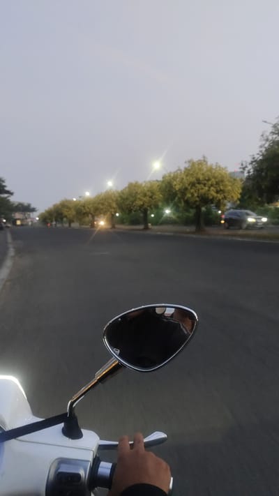 The image is a point-of-view shot from a scooter on a paved road lined with trees in Pusakarakyat, Indonesia. The sky is overcast and light gray, suggesting dusk or dawn. Several streetlights are illuminated along the road, casting a yellow glow. Trees with light green foliage are planted at intervals on the right side of the road, some with their own lights shining on them. A dark-colored car is visible on the right side of the road, its headlights on, and another vehicle is approaching from the distance, its headlights also illuminated. The foreground shows a portion of the white scooter, including the handlebar grip and a hand with dark clothing on the wrist operating the throttle. A chrome rearview mirror is positioned to the right, reflecting the rider and the sky. The road surface is asphalt, and the left side of the road features a sidewalk and some buildings in the far distance.