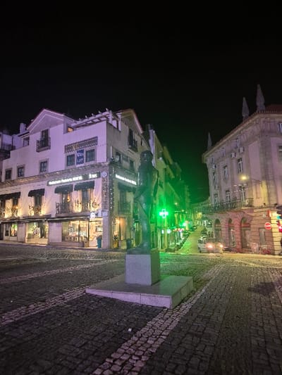 This image presents a nighttime street scene in Covilhã, Portugal, characterized by architectural illumination and artificial light sources.
In the foreground, a bronze-colored statue of a standing male figure is centrally positioned on a two-tiered rectangular grey stone pedestal. The ground is paved with dark cobblestones interspersed with white cobblestone stripes, forming linear patterns.
To the immediate left, a multi-story building with light-colored facades displays "Restaurante Pizzeria HOTEL" and "3.ES" on an illuminated sign at ground level. Its ground-floor windows are lit with warm-toned light, revealing interiors. Balconies with dark railings are visible on upper floors.
To the right, another multi-story building, appearing older with distinct architectural details and multiple windows, features balconies on its upper levels. A red traffic signal is partially visible on the far right.
An uphill street recedes into the background, flanked by buildings. A prominent green illuminated cross, indicative of a pharmacy, is visible on the left side of this street, positioned above other illuminated signs. Further up the street, at least one white van with headlights activated is visible. The sky is uniformly dark, and no human figures are present in the scene.