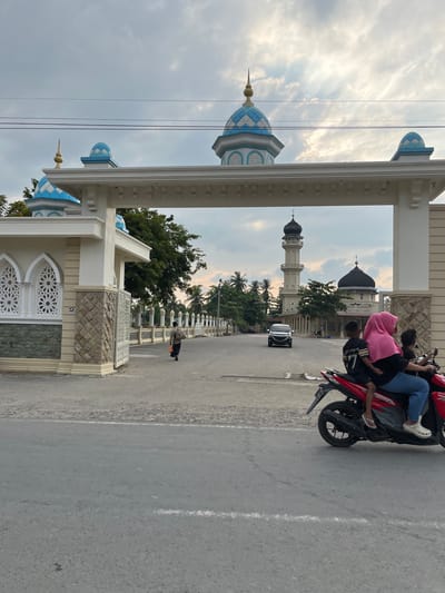 The image captures an entrance to a mosque complex in Dayahbaro, Indonesia, under an overcast sky. A large, white archway, adorned with decorative elements and stone accents, frames the view. Beyond the arch, a minaret and a dome of the mosque are visible. In the foreground, a red motorcycle carries a family of four. The driver, a woman wearing a pink hijab and blue jeans, is in the front, with two children behind her. One child, wearing a black t-shirt with the number "77," sits in front of her, and another child is behind her. A man in dark clothing with a backpack and an orange bag walks away from the motorcycle and towards the mosque entrance. A car is visible driving on the road in the middle ground. The environment appears to be a street leading to a religious site, with palm trees and other vegetation visible in the background.