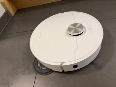 This media file shows a close-up, eye-level shot of a white robot vacuum cleaner on a dark gray tiled floor. The robot vacuum is circular with a metallic, ribbed disc on top, which is likely its charging port or a sensor. There are subtle lines etched into the white surface of the vacuum, suggesting design elements or paneling. To the right of the metallic disc, there are two buttons with icons, one of which has a glowing blue light, possibly indicating it's powered on or in a specific mode. The brand name "DREAME" is visible in gray lettering on the left side of the vacuum's top surface.
The robot vacuum is partially obscuring a dark gray wheel on its underside, which appears to be in motion, indicated by a slight blur. The floor tiles are large and have a matte finish, with visible grout lines. In the upper left corner, a portion of a light-colored wooden edge and a white surface are visible, suggesting the presence of furniture or a wall. The overall scene is indoors, with neutral lighting, and there are no people present. The image is clear and focused on the robot vacuum and its immediate surroundings. The context of Veurne, Belgium, suggests an indoor domestic setting.