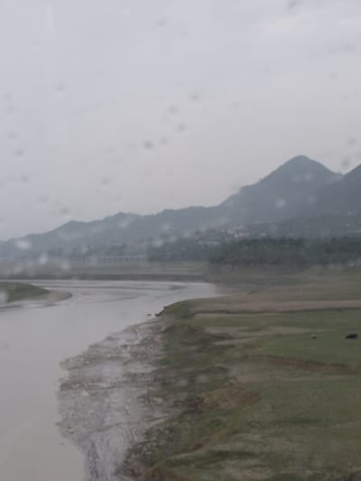 The image captures a landscape scene under an overcast sky, featuring a wide, muddy river flowing in the foreground. The riverbank is uneven and appears to be composed of mud and some sparse vegetation. Beyond the river, a gently sloping, grassy terrain stretches out, dotted with patches of darker earth and vegetation. In the middle ground, a cluster of trees forms a dense line, and behind them, a town or village with buildings is visible nestled at the foot of a mountain range. The mountains themselves are hazy and indistinct due to the atmospheric conditions and distance, their peaks blending with the cloudy sky. The overall impression is one of a rural or semi-rural environment, possibly near a body of water, with a backdrop of rolling hills or mountains. The image appears to have been taken through a window with water droplets on the glass, causing some distortion and obscuring finer details. Without specific landmarks or context, it is not possible to definitively identify the location within Bilaspur, India, but the landscape suggests a region with rivers, hills, and settlements.