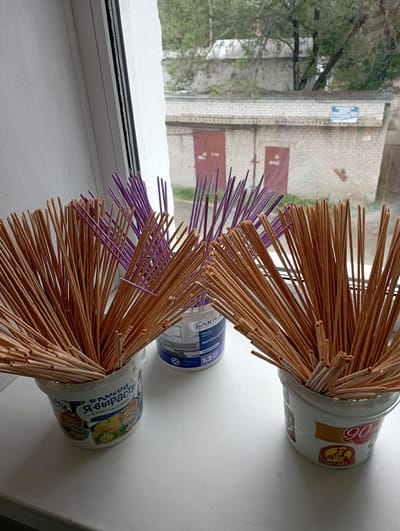 The image depicts a windowsill with three buckets filled with incense sticks. Two buckets contain natural-colored incense sticks, while one bucket contains purple incense sticks. The buckets appear to be repurposed containers, with labels visible on them. One bucket has a label that reads "LAKRA" and "ИНТЕРЬЕРНАЯ" (Interior). Another bucket has a label with the text "#ЯВЫРАСТУ Я БУДУ СЧАСТЛИВЫМ" (I will grow, I will be happy) and cartoon images of bears. The third bucket has a label with the number "90" and the word "КАМЕШК" (kameshok). The background shows a view of buildings and trees through the window, consistent with an urban environment. The presence of garages and brick buildings suggests a residential or industrial area. The overall scene is indoors, looking out onto an outdoor environment. Given the presence of the specific Russian text on the labels, the location is likely Russia. However, without further contextual information or distinctive landmarks, pinpointing the exact location within Blagoveshchensk is not possible.