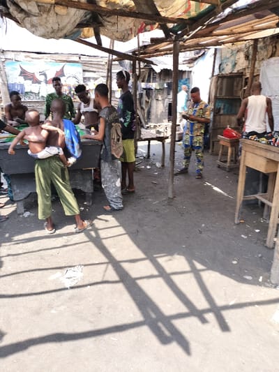 The scene depicts an outdoor gathering under a makeshift shelter in Oworonshoki, Nigeria. The shelter is constructed from rough wooden poles supporting a roof composed of corrugated metal and various patched fabric sheets. The ground is dirt or compacted earth, scattered with debris. Bright sunlight casts distinct, angular shadows from the support structure onto the ground.
Eight individuals are visible. In the foreground, a black rectangular pool table serves as a central point for a group of six males. From left to right around the table: a bare-chested male, a male adolescent in green trousers carrying an infant swaddled in white, a male in a camouflage-patterned shirt, another bare-chested male with a white fabric wrap, a male in grey camouflage trousers carrying a backpack, and a male in a black graphic t-shirt and yellow shorts. The individuals around the pool table appear to be observing or participating in a game.
In the mid-ground, a male wearing a distinctive blue, yellow, and red patterned full-body garment stands, focused on a mobile phone held in both hands. To the right, another male, seen from the back, wears a white tank top and patterned shorts while standing beside a wooden table on which a red object is visible. A light-skinned individual in a light blue shirt is partially visible further in the background, walking away. The background features several dilapidated structures constructed from wood and corrugated metal. A wall on the far left displays a vivid mural of a red and black dragon-like creature.