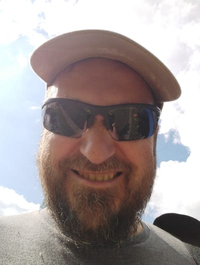 A selfie taken from a low angle shows a man wearing a tan baseball cap and dark sunglasses, looking upwards. The man has a light brown beard and is smiling, with his teeth visible. He is wearing a grey t-shirt. The background is a bright sky with scattered white clouds and some sunlight. The image appears to have been taken outdoors, possibly in a park or open area, given the bright sky and clouds. The location in Ширяево (ильинский), Russia is not discernible from the image itself, as there are no specific landmarks or identifying features of that location.