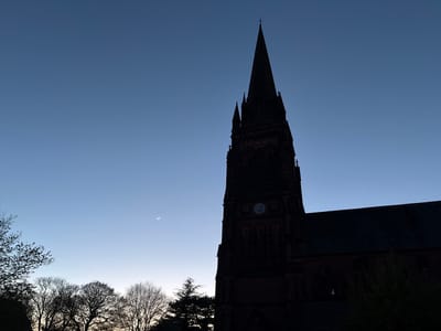The image shows a silhouette of St. Mary's Church in Handbridge, Chester, United Kingdom. The church's spire dominates the right side of the frame. A crescent moon is visible in the blue sky to the left of the spire. Silhouetted trees line the bottom left of the image. The clock on the church tower is visible.