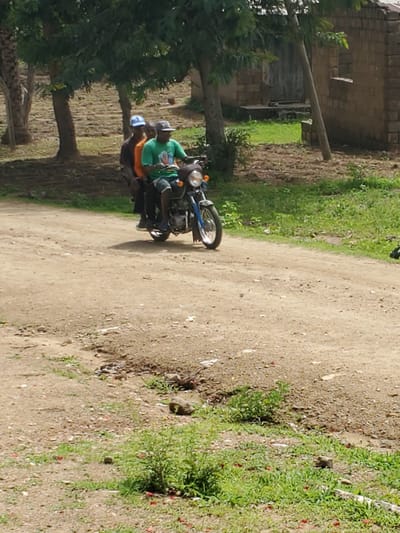 Two people are riding on a motorcycle on a dirt road. The person in front is wearing a blue cap and an orange shirt. The person in the back is wearing a green shirt and a dark cap, and is driving the motorcycle. They are in a rural area, with trees and a brick building in the background. The road is dusty and there is grass on the sides. The location is Kabwir, Nigeria.