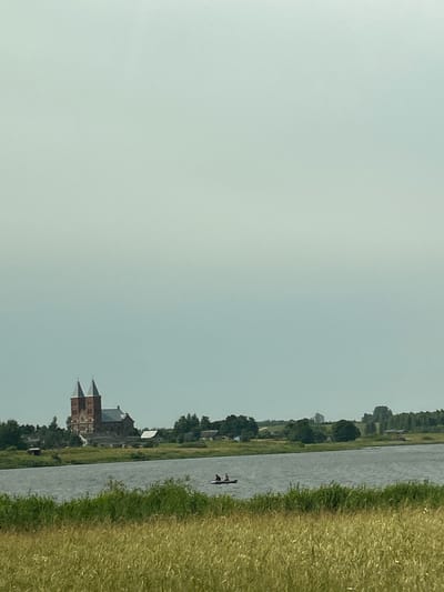 A wide shot captures a serene landscape featuring a river in the foreground, with a village and a large, gothic-style brick church visible across the water. The sky is overcast and pale. In the middle ground, two individuals are paddling a small boat on the river. The foreground is dominated by tall, dry grass, and the middle ground shows lush green vegetation along the riverbank. The background consists of trees and fields, with the prominent church featuring two spires as the focal point of the distant settlement. The overall atmosphere is tranquil and pastoral.