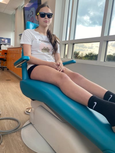 A young female with brown hair pulled back in a ponytail and wearing sunglasses, a white t-shirt with a cat graphic and the word "PURRFEC", and black shorts sits in a bright blue dental chair. She is wearing black Nike socks. The chair is positioned in front of a large window overlooking a treeline and cloudy sky. Behind her, an orange cabinet with drawers is visible. The floor is light-colored wood. A gray cable from dental equipment is visible on the floor next to the chair. The scene appears to be a dental office.