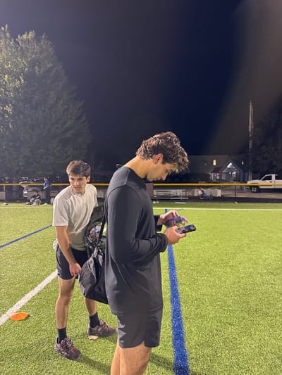 The image depicts two males on an artificial turf sports field at night in Newton, United States. The primary subject, positioned in the foreground on the right, is a male with curly dark hair, wearing a dark long-sleeved athletic shirt and dark shorts. He is engaged with a black smartphone in his left hand and a small purple electronic device in his right, looking downwards at the phone. Slightly behind and to the left of him stands a second male with dark hair, wearing a white short-sleeved t-shirt, dark shorts, and dark athletic shoes. He holds a black duffel bag resting near his left leg and is looking towards the primary subject. The field is green artificial turf marked with white and blue lines; an orange cone lies on the turf in the mid-ground. In the background, beyond a perimeter fence, a treeline, a building with illuminated windows, a flagpole, and a white pickup truck are visible under artificial lighting. Several indistinct figures are present further back on the field.