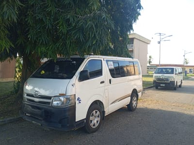 The scene is in Tawpo, Nigeria. A white Toyota HiAce van is parked on a paved road partially under a large, leafy tree. Another white van, bearing the text "MUSLIM STUDENTS," is parked further down the road in the background. Buildings are visible in the background. The environment is an outdoor location, likely a campus or institutional setting, given the presence of the vehicles and buildings.