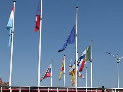 The image displays a collection of flags against a clear blue sky, suggesting a bright, sunny day. The flags are mounted on tall white poles, with one prominent pole on the far left flying a flag with horizontal stripes of light blue, white, and red. To its right, another pole flies a flag that appears to be the flag of Luxembourg, also with horizontal stripes of red, white, and light blue, although it is furled and difficult to discern precisely. Further to the right, a bright blue flag with a circle of yellow stars, the European Union flag, is visible. Other flags are seen at various points in the image, including a flag with red and yellow diagonal stripes (possibly Spain), a flag with red, white, and blue vertical stripes, and a green flag with a white cross. In the lower right of the image, a modern street lamp is visible. In the background, a red-roofed building and a construction crane are partially visible, suggesting an urban or semi-urban setting. The flags themselves appear to be in good condition, gently blowing in the breeze. There is no visible text in the image. The overall impression is one of an international gathering or a location that celebrates diversity through its display of flags, possibly at an event or as a permanent fixture. The scene is static, with the focus on the flags and the sky.