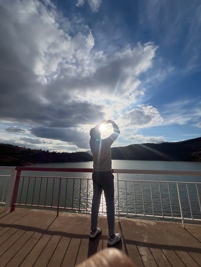 A photograph taken in Tabaroucht, Morocco, shows a person standing on a concrete platform with a metal railing. The person, wearing a light-colored top, grey pants, and white sneakers, is positioned facing away from the camera, holding their hands in a circular shape to frame the sun. The sun is positioned directly behind their hands, causing a bright flare and obscuring the person's silhouette. Behind the person and railing is a body of water, and in the background, a mountainous landscape is visible. The sky is filled with clouds, some of which are partially obscuring the sun. Power lines can be seen stretching across the upper left portion of the sky.