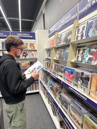 A man with curly hair, wearing glasses, a black hoodie, and green cargo pants, is looking at a vinyl record in a store. He is standing in the "FNAC VINYLE VARIÉTÉ FRANÇAISE" section, surrounded by shelves filled with vinyl records, categorized by sections such as "Variété Internationale". The labels are in French. The location is possibly a FNAC store.