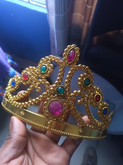 A gold-colored, ornamental crown, adorned with multiple colored jewels, is held by a dark-skinned hand. The crown's embellishments include faux gems in shades of red, green, blue, and pink. Small gold orbs frame the jewels. The band of the crown features a subtle pattern. A backdrop of blurred textures includes curtains, a window, and other indistinct objects.