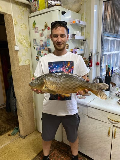 A man is holding a large fish in a kitchen. The man is wearing shorts and a t-shirt with a design. A refrigerator with various items on it is visible in the background, along with a window and kitchen cabinets. The floor is carpeted. The location is Баклуши, Russia.