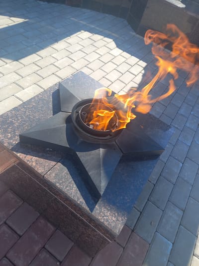 A close-up, high-angle view shows an eternal flame in a memorial setting. The flame burns brightly in a dark metal receptacle, which is set atop a black, granite-like star-shaped monument. The monument is surrounded by paving stones, some light grey and others a reddish-brown. Shadows from an unseen object stretch across the grey paving stones. In the background, darker stone structures, possibly part of a larger monument or building, are visible. The scene suggests a solemn and commemorative location, likely in a public space within the city of Sterlibashevo, Russia.