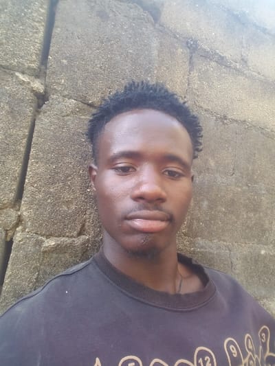 A young Black man poses against a stone wall. He wears a dark T-shirt with a light-colored graphic on the left side. His hair is short and dark. He faces slightly to the right, looking directly at the camera with a neutral expression. The lighting highlights his face and the texture of the wall behind him.