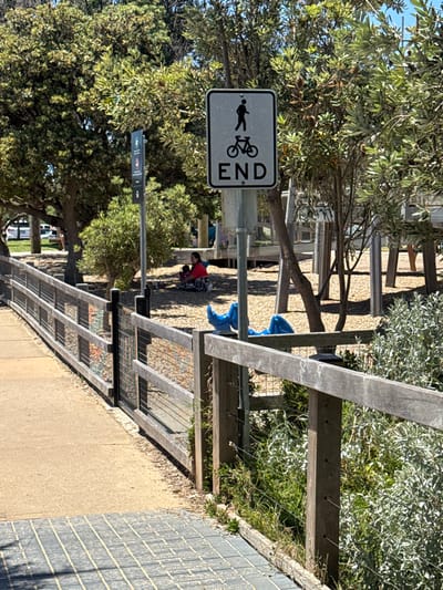 The image depicts an outdoor recreational area or shared pathway under bright, clear daylight in Frankston, Australia. A paved path or sidewalk runs along the bottom and left side of the frame, leading to a gravel area further into the scene.

A prominent white rectangular sign with a black border is positioned on the left side of the path. It features black silhouettes of a pedestrian and a bicycle symbol, with the word "END" written vertically below them, indicating the conclusion of a shared path for walkers and cyclists.

Along the right side of the path, a sturdy wooden fence or barrier, constructed with horizontal wooden beams and sections of wire mesh, separates the pathway from an area dense with green foliage and bushes. Further in the background, near the gravel path, a person wearing a red top and patterned pants is visible, appearing to be crouched or seated on the ground. Two bright blue objects, possibly bags, are located close to this person. The area is well-treed, with numerous trees featuring lush green leaves providing shade throughout the scene. The clear lighting suggests a sunny day.