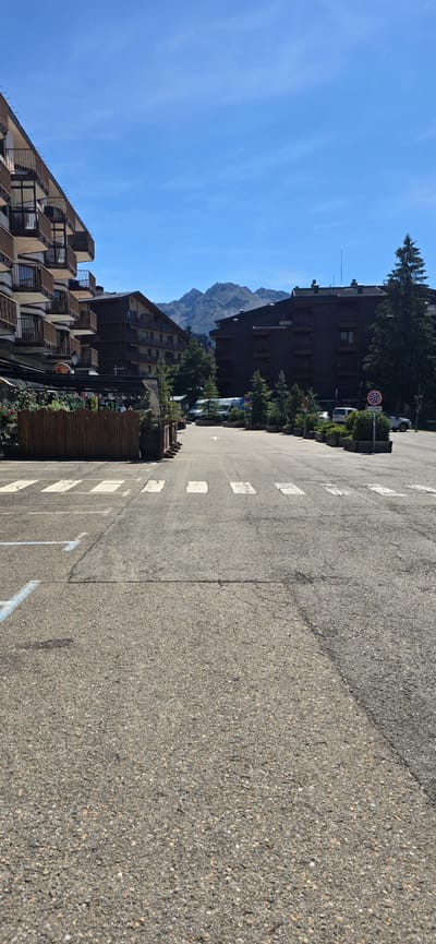 The image captures a street scene in Sallent de Gállego, Spain, under a clear blue sky with distant mountains. The foreground features a paved road with a pedestrian crossing marked by white stripes. To the left, a multi-story building with numerous balconies is visible, adorned with dark brown railings. Adjacent to this building is a wooden fence enclosing an outdoor seating area, likely a restaurant or cafe, with greenery and tables. Across the street, more multi-story buildings with dark facades and balconies are present, along with several evergreen trees. Parked vehicles, including a white van and a car, are visible on the right side of the road. A circular sign with red and blue symbols indicating a restriction is also situated on the right. The overall scene depicts a tranquil, daytime setting in a mountainous town.