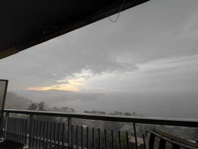 This is a photograph taken from a balcony overlooking a coastal city. The sky is overcast and grey, with a band of lighter clouds and a hint of yellow light on the horizon suggesting either dawn or dusk. In the distance, mountains are visible through a haze or fog. The city below appears to be built on hills, with buildings clustered together. A body of water, possibly a bay or gulf, is visible in the middle ground, with some structures or ships on it. The foreground shows the railing of the balcony, made of metal bars, and a cushioned chair. The overall atmosphere is somber and slightly melancholic, possibly due to the weather conditions. The image depicts a view in the Municipal Unit of Aegio, Greece, characterized by its hilly terrain and proximity to the sea.