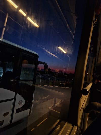 The image captures a view from inside a bus at dusk, with the sky displaying hues of deep blue and pink. The bus's interior is visible, including seats and overhead lights, some of which are illuminated. Reflections on the bus windows show lights from outside and the twilight sky. Outside the bus, a parking lot or roadside area is visible with several parked cars. A fence separates the road from a grassy area. The overall scene suggests a stop or a journey during evening hours. The location is identified as Bahovitsa, Bulgaria.