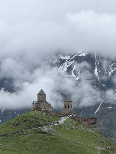 The image depicts the Gergeti Trinity Church, a 14th-century Orthodox church located in the village of Gergeti, Georgia. The church is situated on a prominent hilltop, surrounded by the majestic Caucasus Mountains, which are partially obscured by thick, swirling clouds and patches of snow. The architecture of the church is characterized by its stone construction, a central dome topped with a cross, and an adjacent bell tower. A pathway winds its way up the grassy hillside towards the church, and several small figures of tourists can be seen walking along it, exploring the grounds, and admiring the view. The overall atmosphere is one of dramatic natural beauty and historical significance, with the rugged mountain landscape and the ancient church creating a powerful visual.