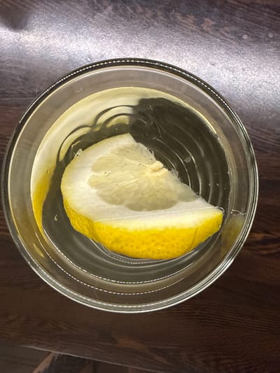 A close-up, overhead shot captures a single slice of lemon floating in a clear liquid within a glass. The lemon slice, bright yellow on its rind and pale yellow and white on its flesh, is partially submerged. The liquid is clear with subtle ripples and a few small air bubbles, suggesting it is water or a similar beverage. The glass has a thick rim with a decorative, dotted line pattern. The glass sits on a dark, wooden surface with a subtle grain texture, and a lighter, metallic-like surface is visible in the upper portion of the frame. There are no people or any other objects visible in the scene. The image appears to be taken indoors in a dimly lit environment. There is no information to ascertain its location in Kondopoga, Russia.