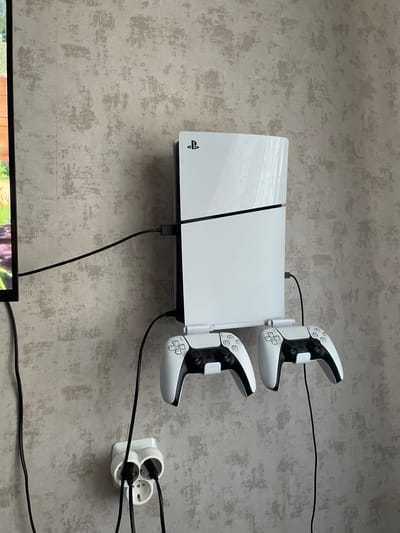 A white PlayStation 5 console is mounted horizontally on a textured gray wall, with two white PlayStation 5 controllers attached to a charging dock mounted on the side of the console. The controllers are oriented vertically, one above the other, with their grips facing downwards and their joysticks and buttons facing the viewer. The console has the PlayStation logo on its left side. A black cable extends from the top of the console and connects to a white triple power adapter plugged into the wall. Two other black cables also run from the power adapter, presumably to power the console and potentially other devices. The setting appears to be indoors, likely a living room or entertainment area, given the presence of a television screen visible at the bottom of the frame. The lighting suggests it is daytime, and there are no overt weather cues. The scene is neatly organized, with the gaming equipment mounted to save space and keep it accessible.