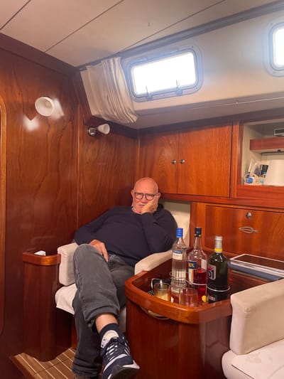 An older male subject is seated within the polished wooden interior of a yacht cabin, located in La Grande-Motte, France. The man, bald with a white mustache and wearing black-framed glasses, a dark blue long-sleeved shirt, and dark grey jeans, occupies a light-colored armchair. His right hand supports his chin, and his left leg is crossed over his right, revealing a dark blue and white sneaker.
The cabin walls and built-in cabinetry are constructed from highly polished reddish-brown wood. Two rectangular portlight windows are visible on the upper wall, with a partially drawn beige curtain over the left window. Two wall-mounted light fixtures with white conical shades are affixed to the left wall.
To the subject's right, a small, polished wooden table is positioned between two armchairs. On this table, three distinct bottles of alcohol are arranged in integrated holders: a bottle of Santalu Duo (clear spirit), a bottle of Aperol (orange-red liqueur), and a bottle of Pastis 51 (yellow-green label). Two clear drinking glasses are also present, one containing an amber liquid with ice, the other appearing empty. A pair of dark sunglasses rests near the glasses. The wooden cabinetry includes various compartments with small silver handles and an open shelf displaying miscellaneous items to the far right. A section of wooden slatted flooring is visible beneath the man's feet.