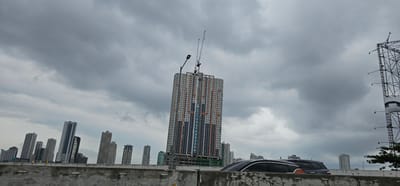 This image captures a view of the Manila skyline under a cloudy sky. The dominant feature is a tall, multi-story building under construction, adorned with orange, white, and dark gray panels. Two construction cranes are positioned at the top of this building. A light pole stands next to the building. To the left of the construction site, several other high-rise buildings of varying heights and architectural styles form part of the cityscape. In the foreground, a concrete barrier runs horizontally across the frame. A dark-colored SUV is partially visible driving on a road behind the barrier. To the far right of the image, a large billboard structure is visible. The overall scene suggests an urban environment in Manila, Philippines, with ongoing development.