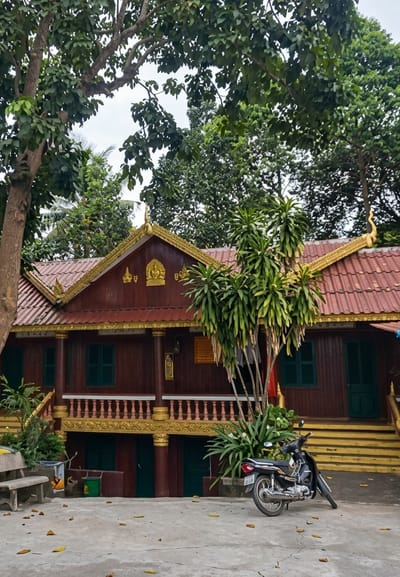 This is a full-frame, eye-level, outdoor, vertical shot of a Buddhist temple in Sihanoukville, Cambodia. The temple has a traditional wooden structure with a dark red exterior and a red tiled roof. The roof is adorned with ornate golden trim and decorative elements. Green shutters are visible on the windows. A balcony with golden railings runs along the front of the temple. A motorcycle is parked on the concrete ground in front of the temple, to the right of a cluster of green plants. A stone bench is situated to the left of the plants. Yellow stairs lead up to the right side of the temple. Trees with lush green foliage surround the temple. The ground is a concrete patio with fallen leaves scattered across it. The sky is overcast.