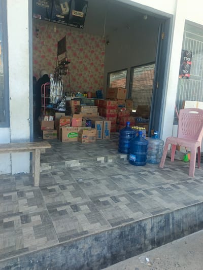 The image shows the exterior of a small convenience store in Tenggela, Indonesia. In the foreground, a grey tiled floor with a geometric pattern is visible, leading up to a raised concrete step. To the left, a simple wooden bench with two legs sits on the tiles.
The store entrance is open, revealing a cluttered interior. Boxes of various goods are stacked inside and in front of the entrance. Among the visible items are boxes labeled "Kompas," "Super," and "Lemonia." Several large blue water jugs are placed on the tiled floor just outside the entrance, with one clear jug also present.
To the right of the entrance, a pink plastic chair is positioned. Behind the chair, a broom with green and orange bristles leans against the wall. On the wall to the right of the entrance, two circular posters with the word "TROY" are displayed.
Inside the store, a person wearing a dark jacket and a white helmet is partially visible, appearing to be working or attending to something. A metal shelf unit holding more products is also seen within the store. The interior walls are decorated with a floral wallpaper featuring pink roses.
The overall scene suggests a typical small, local business in Indonesia, with a focus on selling various household goods and beverages. The presence of water jugs indicates they are likely a distributor or seller of bottled water.