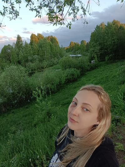 The image is a selfie taken outdoors in a rural area. A fair-skinned woman with blonde hair is in the foreground, looking at the camera. She has a noticeable bruise around her left eye. She is wearing a dark-colored hoodie with a white pattern on the front. The background consists of a green, grassy hillside. Beyond the hill, there is a wooded area with various green trees and bushes. A small body of water, possibly a pond or stream, is visible among the trees. The sky above the trees is light with some clouds, suggesting it might be dusk or dawn. Based on the information provided, this scene is in Verkhnii Dvor, Russia. There are no specific elements from the photograph that would confirm the location other than the provided text.