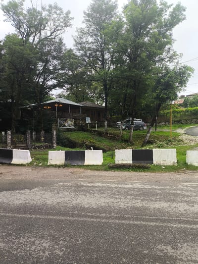 The image captures a street-level view of a roadside establishment in Gachedili, Georgia. In the foreground, a paved road with visible water patches is bordered by a row of black and white concrete barriers. Behind these barriers, a grassy slope leads up to a rustic building complex. This complex features a covered outdoor dining area with lights and a traditional-looking building behind it. A metal fence with ornate posts separates the property from the road. Several trees, some quite large, surround the establishment, creating a lush, green environment. Two cars are parked on the property behind the fence. The sky is overcast, suggesting a cloudy day.