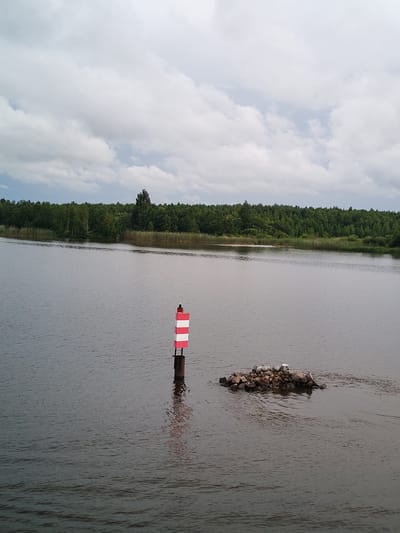 A serene, wide shot captures a tranquil body of water, likely a river or lake, on an overcast day. The water's surface is calm, with gentle ripples disturbing its dark, reflective expanse. On the left side of the frame, a navigational buoy stands tall, its red and white stripes clearly visible, marking the waterway. To the right of the buoy, a small, rocky island emerges from the water, appearing to be artificially constructed or a natural formation. A single white object, possibly a bird or debris, rests atop the island.
The background is dominated by a lush, dense forest that stretches across the horizon, its green canopy a stark contrast to the muted tones of the sky and water. The trees appear to be a mix of deciduous and coniferous species, with a line of taller reeds or marsh grass visible along the water's edge, separating the water from the forest. The sky above is filled with heavy, grey clouds, suggesting a possibility of rain or simply a cloudy day. There are no people or animals visible in the image, apart from the single white object on the island. The scene evokes a sense of natural beauty and peacefulness, with the man-made elements like the buoy and the island integrated into the landscape.
This location is identified as Kargulino, Russia.