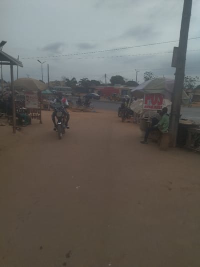 The image shows a street scene in Junction, Nigeria (latitude: 8.958, longitude: 7.551). The scene is taken from a slight elevation on a dirt road, looking towards the street. The sky is overcast with a hint of blue, with the sun obscured. There are trees lining the street, and buildings in the distance. A man is riding a motorcycle on the right side of the road, heading toward the viewer. There are two makeshift stalls in the foreground, both labeled with signs that read "POS AVAILABLE," suggesting they are selling goods or providing services. Several people are standing or sitting in the vicinity, with a few motorcycles parked near the stalls. The ground is a light brown dirt, and there are some scattered pieces of trash on the ground. The overall atmosphere is somewhat dusty and quiet. The scene appears to be a typical street scene in Junction, with a mix of street vendors, locals, and motorcycles, which is common in the area.