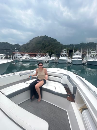 A young woman wearing a light brown halter top, black skirt, and sunglasses sits on a white boat in the marina of Playa Herradura, Costa Rica. The woman is holding a blue and silver can in her left hand and has her right hand resting on the boat’s seat. Behind her are numerous boats docked in the marina, all facing the same direction. The woman is smiling and looking directly at the camera. The marina is lined with palm trees and greenery. The sky is mostly cloudy and overcast. The water in the marina is a clear blue-green color. The image was taken from the interior of the boat. The woman is the only person visible in the image. The coordinates (9.651, -84.665) place the image in the marina of Playa Herradura, Costa Rica.