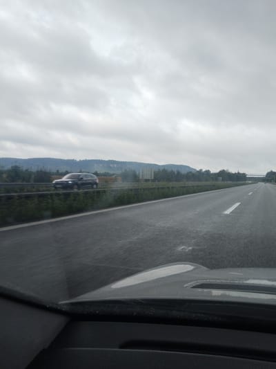 This is a photograph taken from the perspective of a vehicle traveling on a highway, likely in or near Loukov, Czechia, given the context provided. The sky is overcast with a dense layer of clouds, suggesting a dull or possibly rainy day. The highway itself appears to be wet, with reflections visible on the asphalt, indicating recent precipitation or ongoing light rain. White lane markings are visible on the road.

To the left of the highway, a metal guardrail separates the road from a grassy median and a row of trees. Beyond the trees, rolling hills or mountains are visible, covered in a mix of trees and possibly some fields or cleared areas. A dark-colored SUV is captured in motion on the highway to the left, appearing blurred due to the vehicle's movement and the camera's perspective. The vehicle is in the left lane of the highway. In the distance, a highway overpass or bridge can be seen crossing the road, with some distant lights visible beneath it. On the left side of the highway, further in the distance, there are some large, light-colored rectangular signs. The foreground of the image shows the dashboard and hood of the vehicle from which the photo was taken.