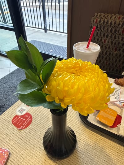Here is a detailed description of the image:
**Scene:** The image appears to be taken from an overhead perspective on a table within an establishment in Stone Mountain, Georgia, United States. The scene features a table set for dining.
**Objects:**
* A vase containing a large, bright yellow chrysanthemum flower.
* A drink cup with a red straw is visible.
* A Chick-fil-A food tray with takeout items, including a Honey Mustard packet.
* A QR code with the message "Order from the comfort of your table" with the information that you scan the code, place the order, and it will be brought to the table.
* A small patterned purse.
**Environment:**
* The table has a textured surface.
* A window or opening with a view of an exterior sidewalk is visible.
* Seating with patterned upholstery is seen in the background.
**Overall Impression:** The image provides a snapshot of a casual dining experience within an establishment that offers table ordering.