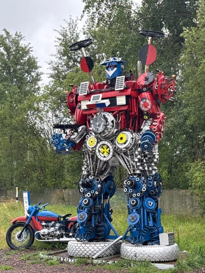 This image shows a large, red and blue robot sculpture made from recycled metal parts, situated outdoors in what appears to be a rural or park-like setting. The robot is detailed, with visible gears, pipes, and other mechanical components. It stands tall, with solar panels on its chest and what looks like a steering wheel and a saw blade attached to its head. To the left of the robot is a vintage blue and red motorcycle. The background consists of lush green trees and a cloudy sky. A concrete barrier runs horizontally behind the motorcycle and robot. There is also a small white sign with some text in a foreign language near the motorcycle. The ground is covered with gravel and grass, suggesting an outdoor exhibition or a roadside attraction. The location is specified as Палыкавіцкі сельскі Савет, Belarus.