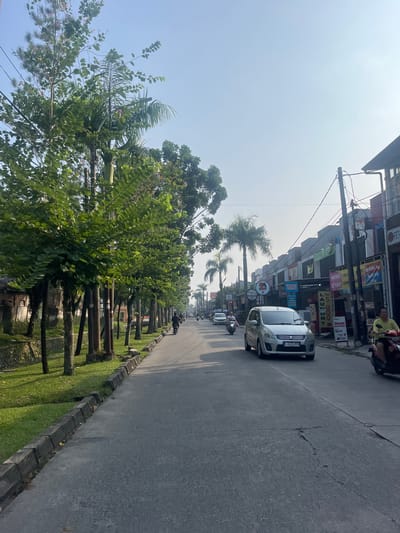 The image depicts a street scene in Ciakar, Indonesia, characterized by a paved road lined with trees on the left and commercial buildings on the right. The road has two lanes and shows some wear with visible cracks. Several vehicles, including cars and motorcycles, are traveling on the road. On the left side of the road, there is a grassy area with a row of trees and a stone retaining wall. The buildings on the right appear to be shops with various signs and advertisements, suggesting a commercial district. The sky is clear and blue, indicating fair weather. A few individuals are visible on motorcycles, and one person is seated near a building. The overall impression is a typical street view in a populated area of Ciakar.